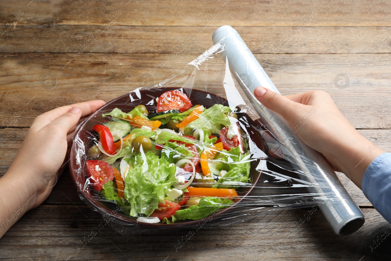 Photo of Woman putting plastic food wrap over bowl of fresh salad at wooden table, closeup