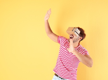 Young man using cardboard virtual reality headset on color background. Space for text