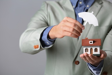 Photo of Male agent covering house model with umbrella cutout on grey background, closeup. Home insurance