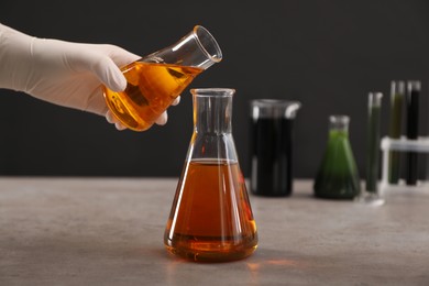 Photo of Woman pouring yellow crude oil into flask at grey table against dark background, closeup