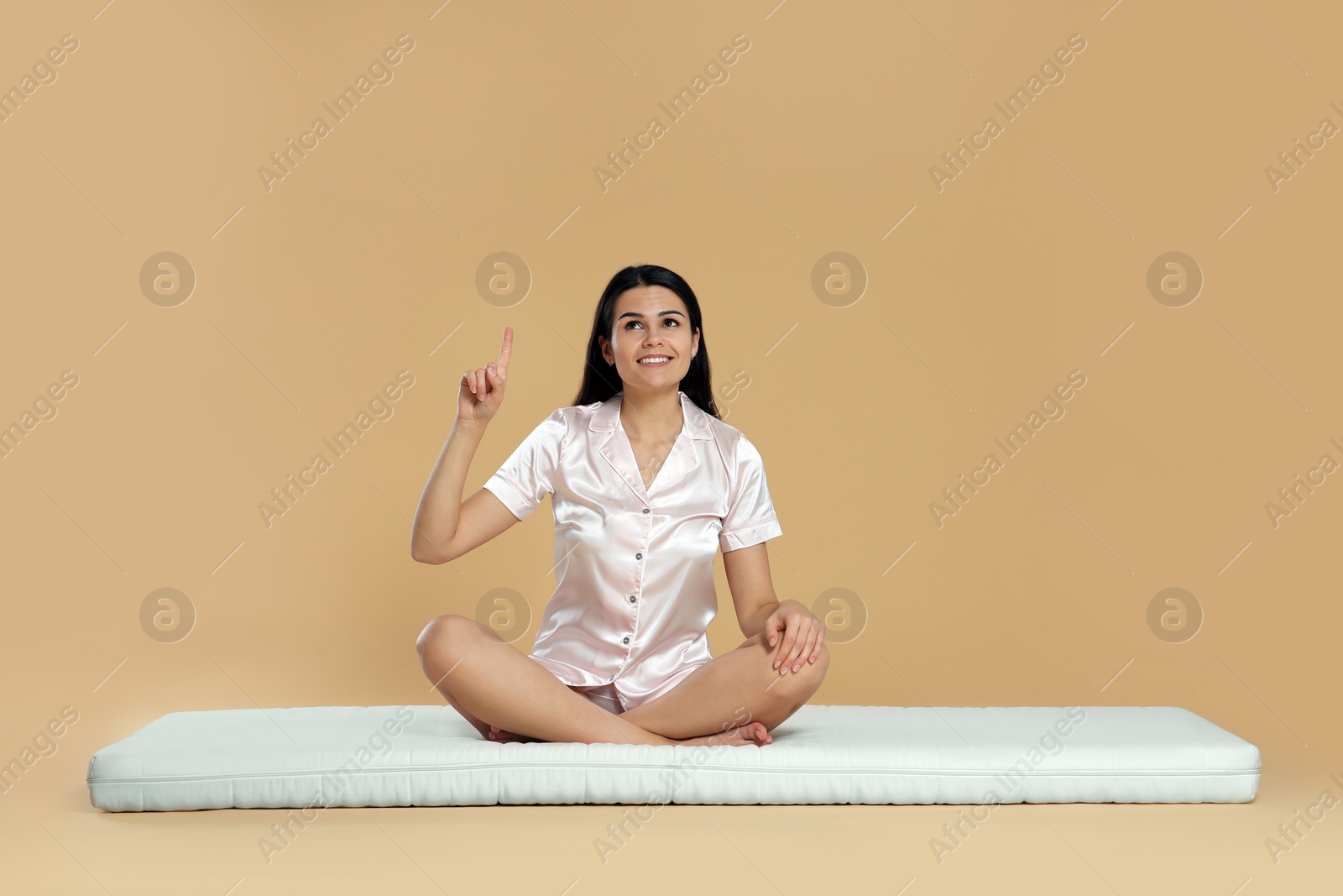 Photo of Young woman sitting on soft mattress and pointing upwards against beige background