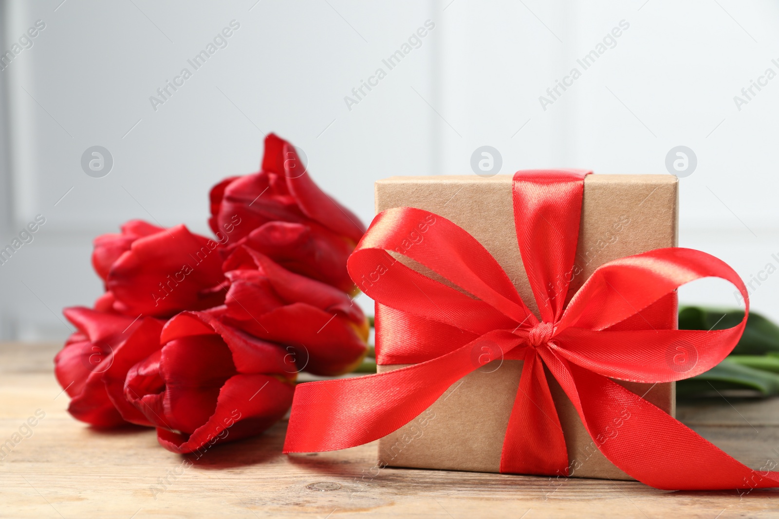 Photo of Beautiful gift box with bow and red tulips on wooden table, closeup