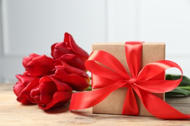 Beautiful gift box with bow and red tulips on wooden table, closeup