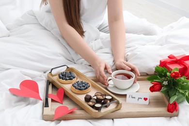 Photo of Tasty breakfast served in bed. Woman with tea, desserts, gift box, flowers and I Love You card at home, closeup