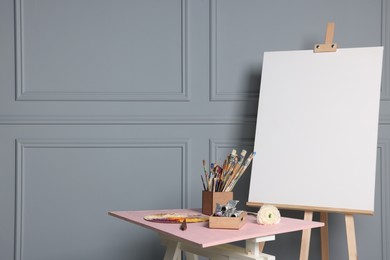 Photo of Easel with blank canvas and different art supplies on wooden table near grey wall. Space for text