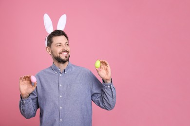 Happy man in bunny ears headband holding painted Easter eggs on pink background. Space for text