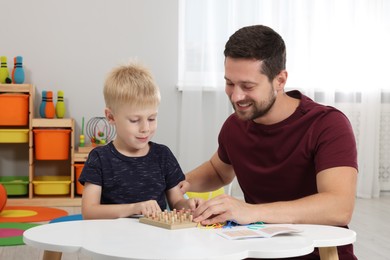 Photo of Motor skills development. Happy father helping his son to play with geoboard and rubber bands at white table in room