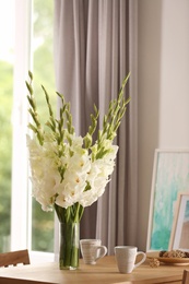Photo of Vase with beautiful white gladiolus flowers, pictures and cups on wooden table in room, space for text