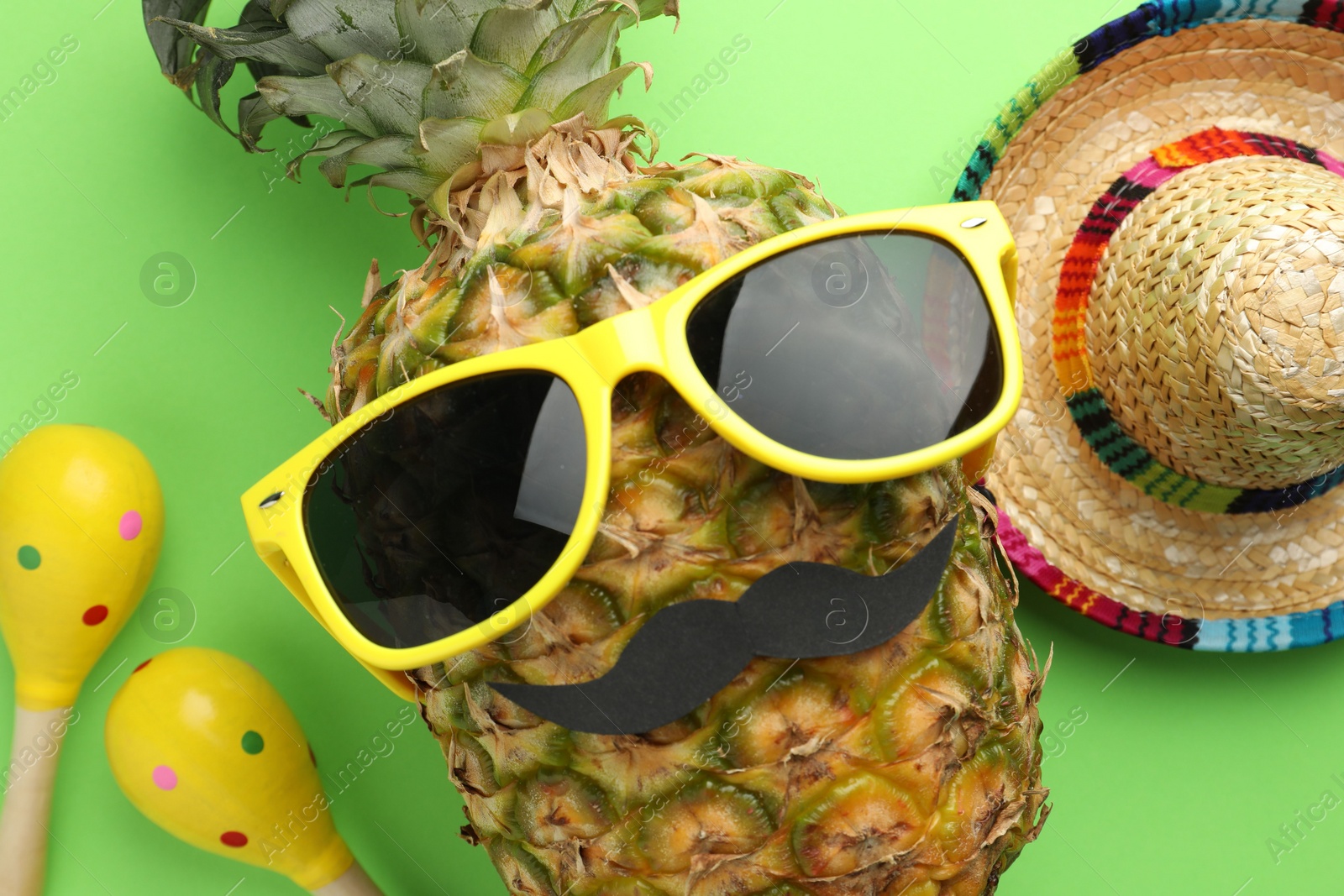 Photo of Mexican sombrero hat, pineapple with sunglasses, fake mustache and maracas on green background, flat lay