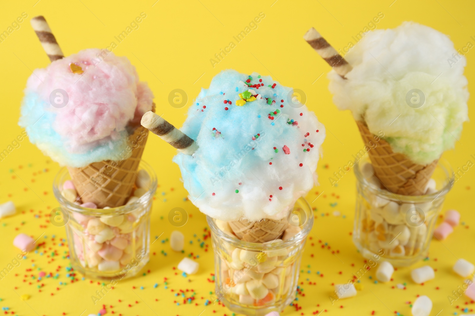 Photo of Sweet cotton candies in waffle cones and marshmallows on yellow background, closeup