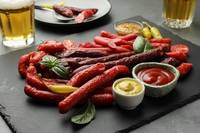 Photo of Different thin dry smoked sausages, sauces and glasses of beer on grey table