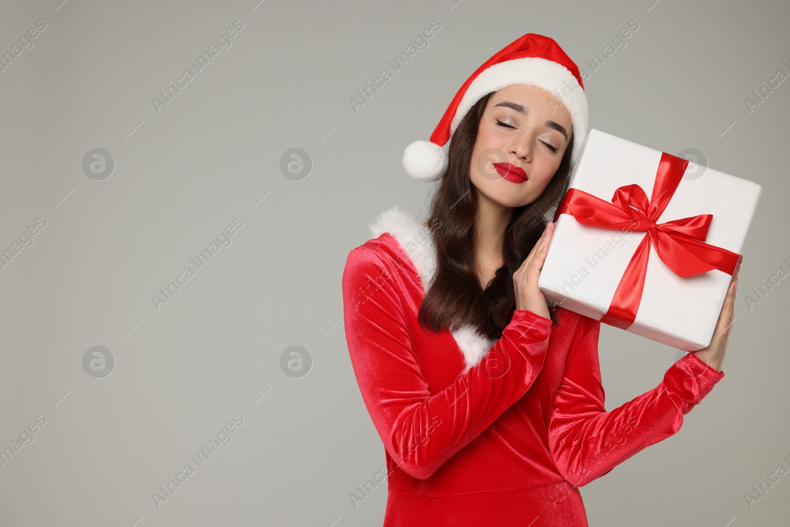 Photo of Beautiful young woman in red dress and Santa hat with Christmas gift on grey background, space for text