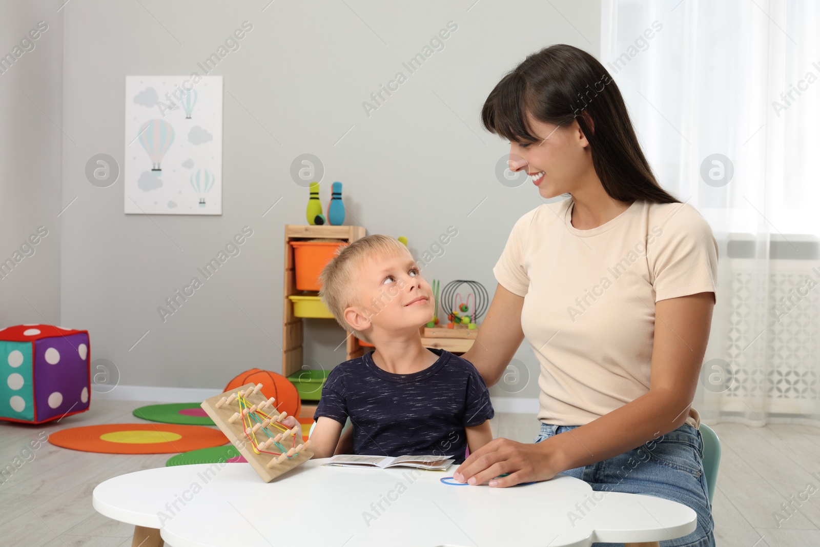 Photo of Motor skills development. Happy mother helping her son to play with geoboard and rubber bands at white table in room