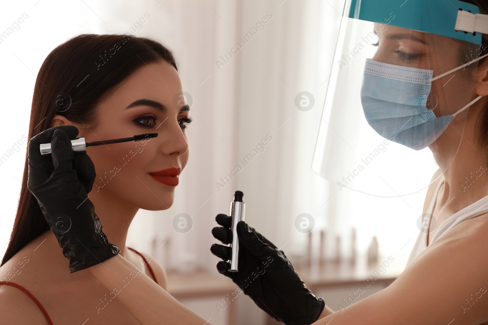 Photo of Makeup artist in protective mask and gloves working with woman indoors. Preventive measures during COVID-19 pandemic