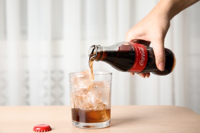 MYKOLAIV, UKRAINE - NOVEMBER 15, 2018: Woman pouring Coca Cola into glass with ice cubes at table, closeup