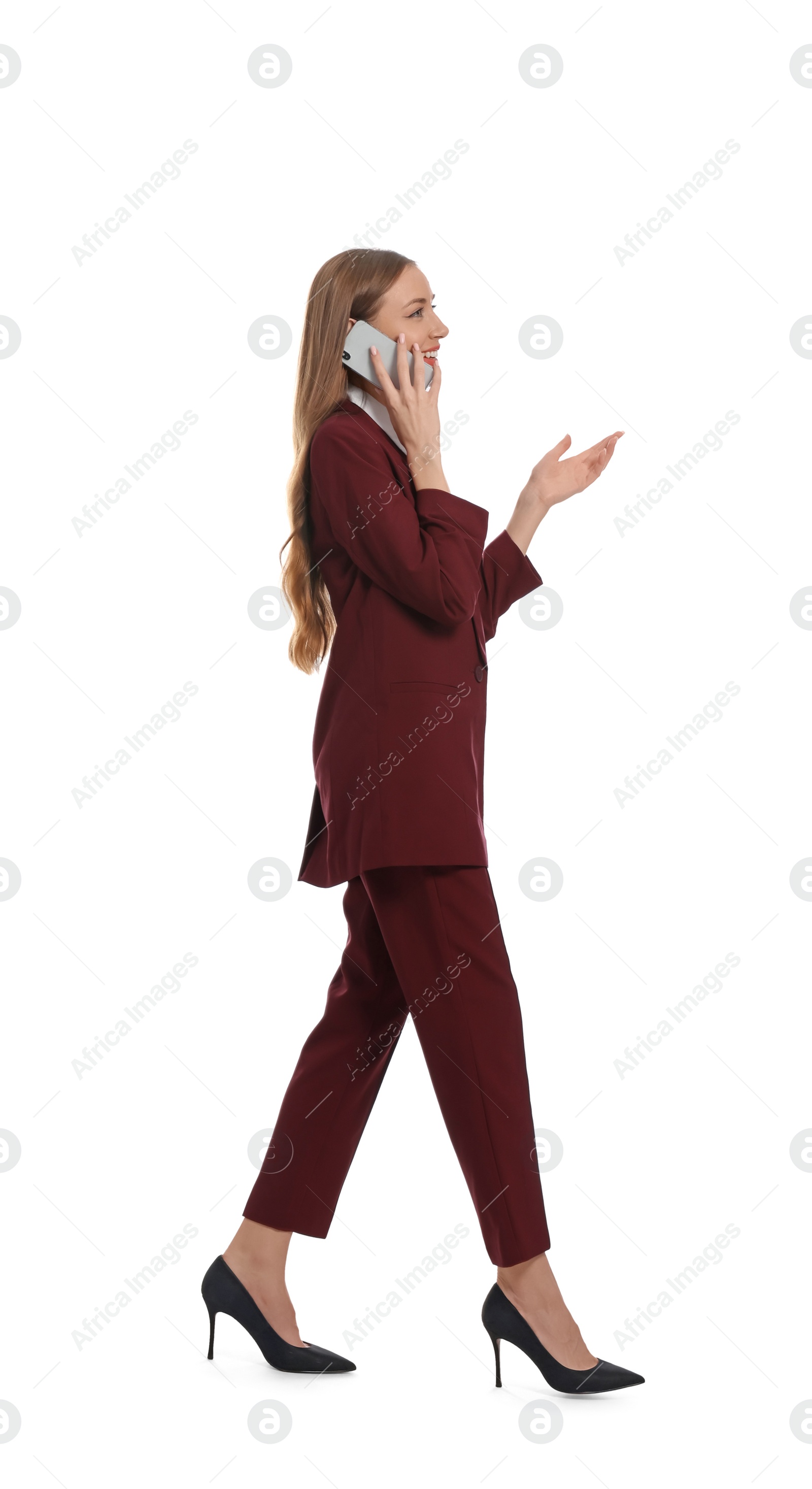 Photo of Young woman in burgundy suit talking on smartphone while walking against white background