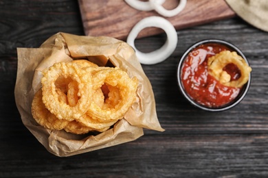 Dishware with homemade crunchy fried onion rings and tomato sauce on wooden table, top view