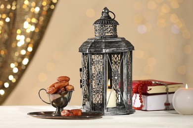 Photo of Arabic lantern, Quran, misbaha, candle and dates on white table against blurred lights
