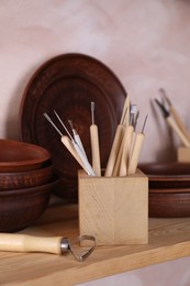 Photo of Set of different crafting tools and clay dishes on wooden rack in workshop