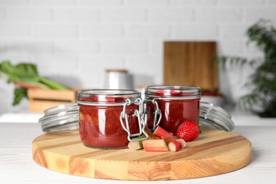 Jars of tasty rhubarb jam, cut stems and strawberry on white wooden table