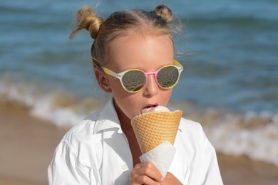 Photo of Adorable little girl eating delicious ice cream near sea on sunny summer day