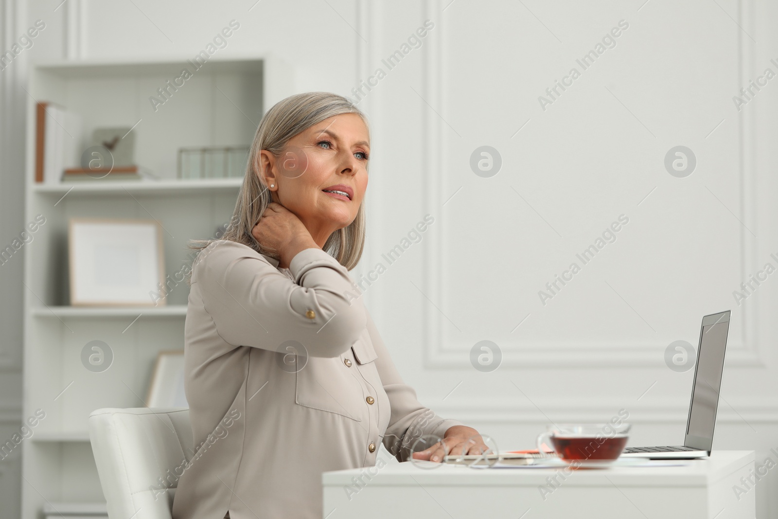 Photo of Woman suffering from neck pain at workplace in room