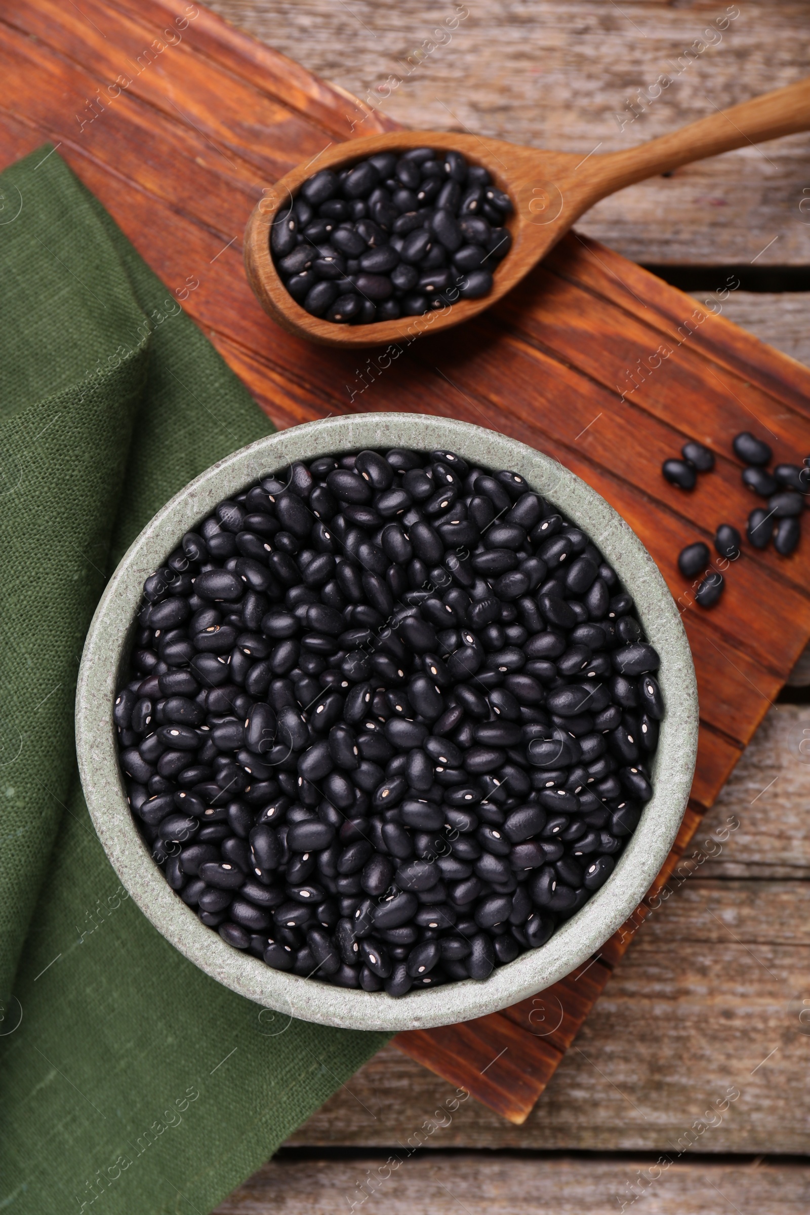 Photo of Bowl and spoon of raw black beans on wooden table, flat lay
