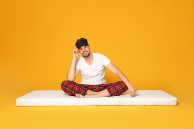 Photo of Smiling man in sleeping mask sitting on soft mattress against orange background