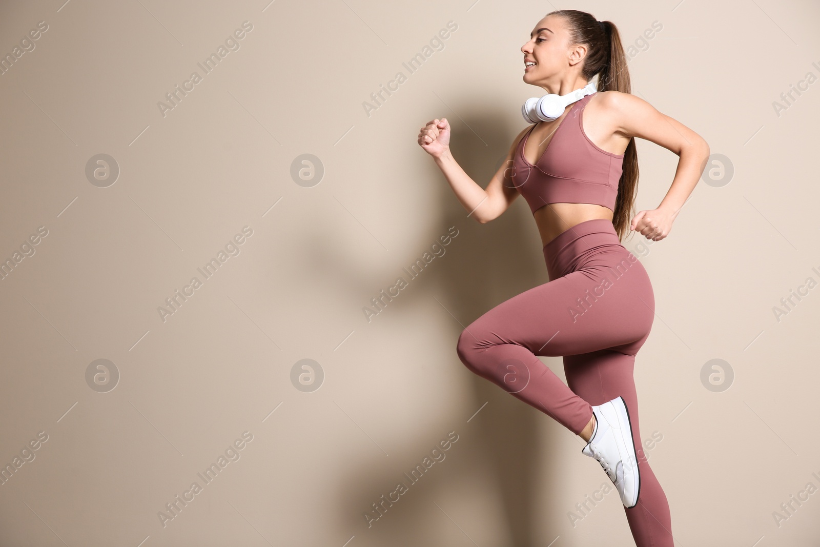 Photo of Young woman in sportswear with headphones running on beige background. Space for text