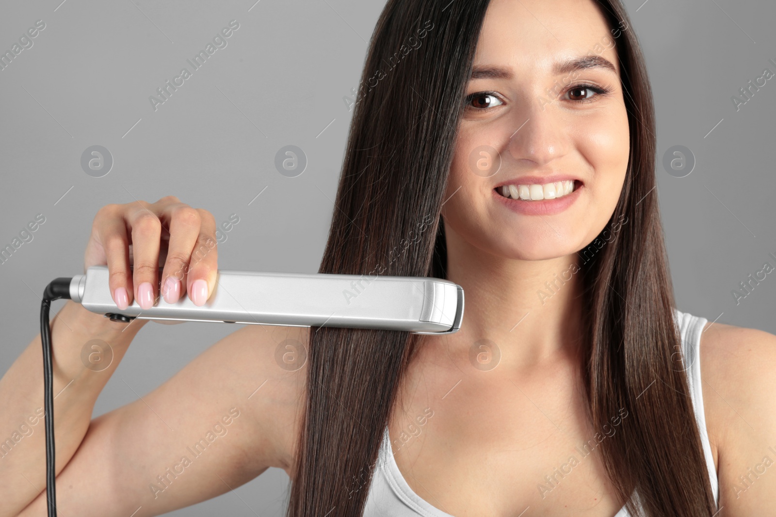 Photo of Young woman using hair iron on grey background