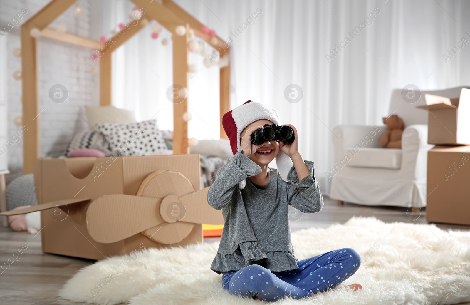 Photo of Cute little girl playing with binoculars and cardboard airplane in bedroom