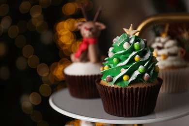 Photo of Christmas tree shaped cupcake on white stand against blurred festive lights, space for text