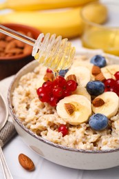 Honey pouring into bowl of oatmeal with berries, almonds and banana slices on white wooden table, closeup