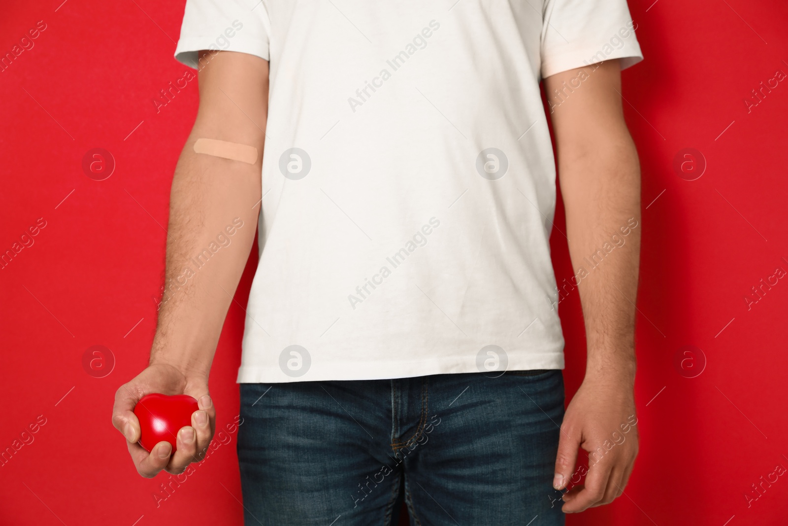 Photo of Blood donation concept. Man with adhesive plaster on arm holding red heart against color background, closeup