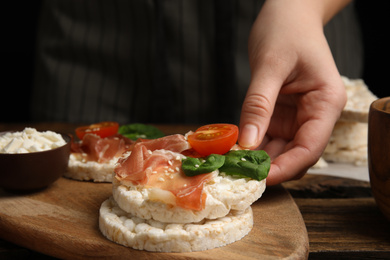 Photo of Woman holding puffed rice cake with prosciutto, tomato and basil at wooden table, closeup