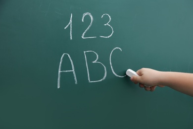 Photo of Little child writing letters and numbers on chalkboard, closeup