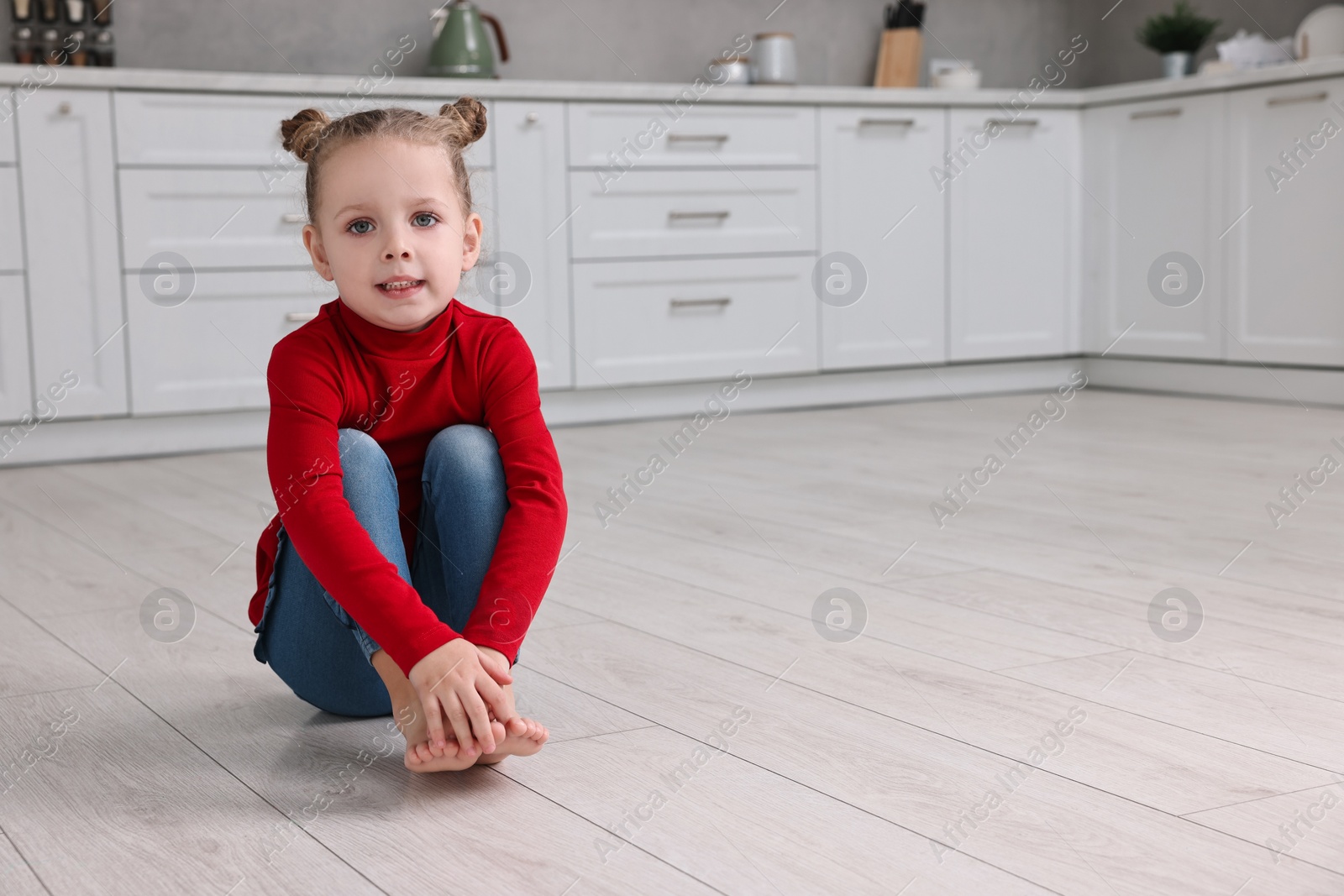 Photo of Cute little girl sitting on warm floor in kitchen, space for text. Heating system
