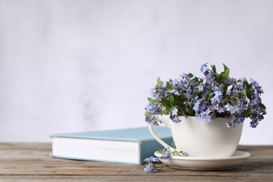 Photo of Beautiful forget-me-not flowers in cup, saucer and book on wooden table against light background, closeup. Space for text