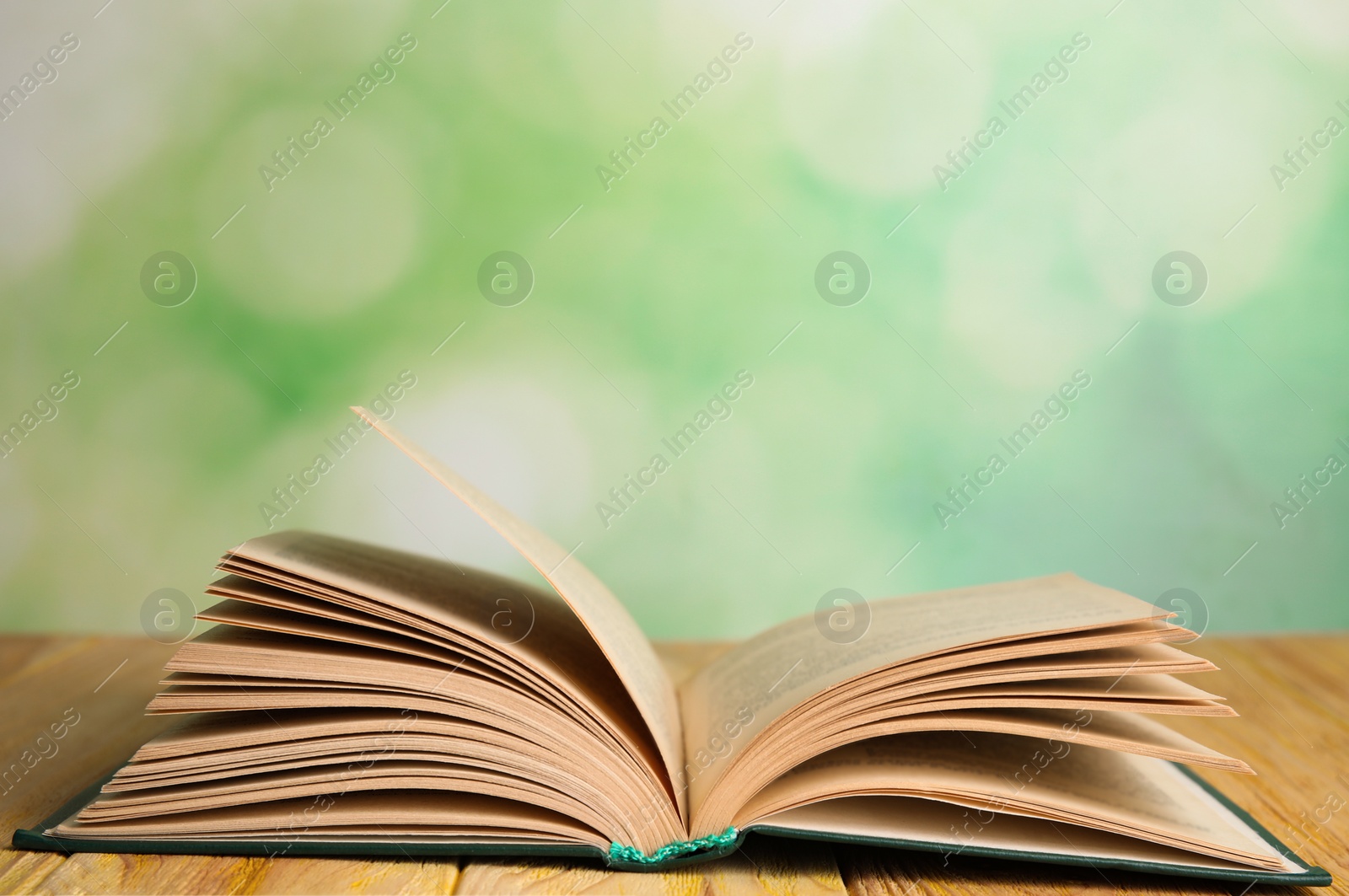 Photo of Open book on wooden table against blurred green background. Space for text