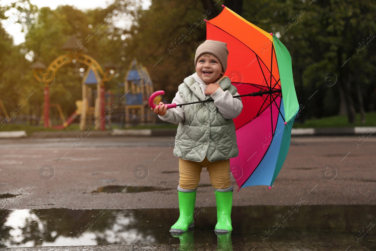 Photo of Cute little girl with colorful umbrella standing in puddle outdoors