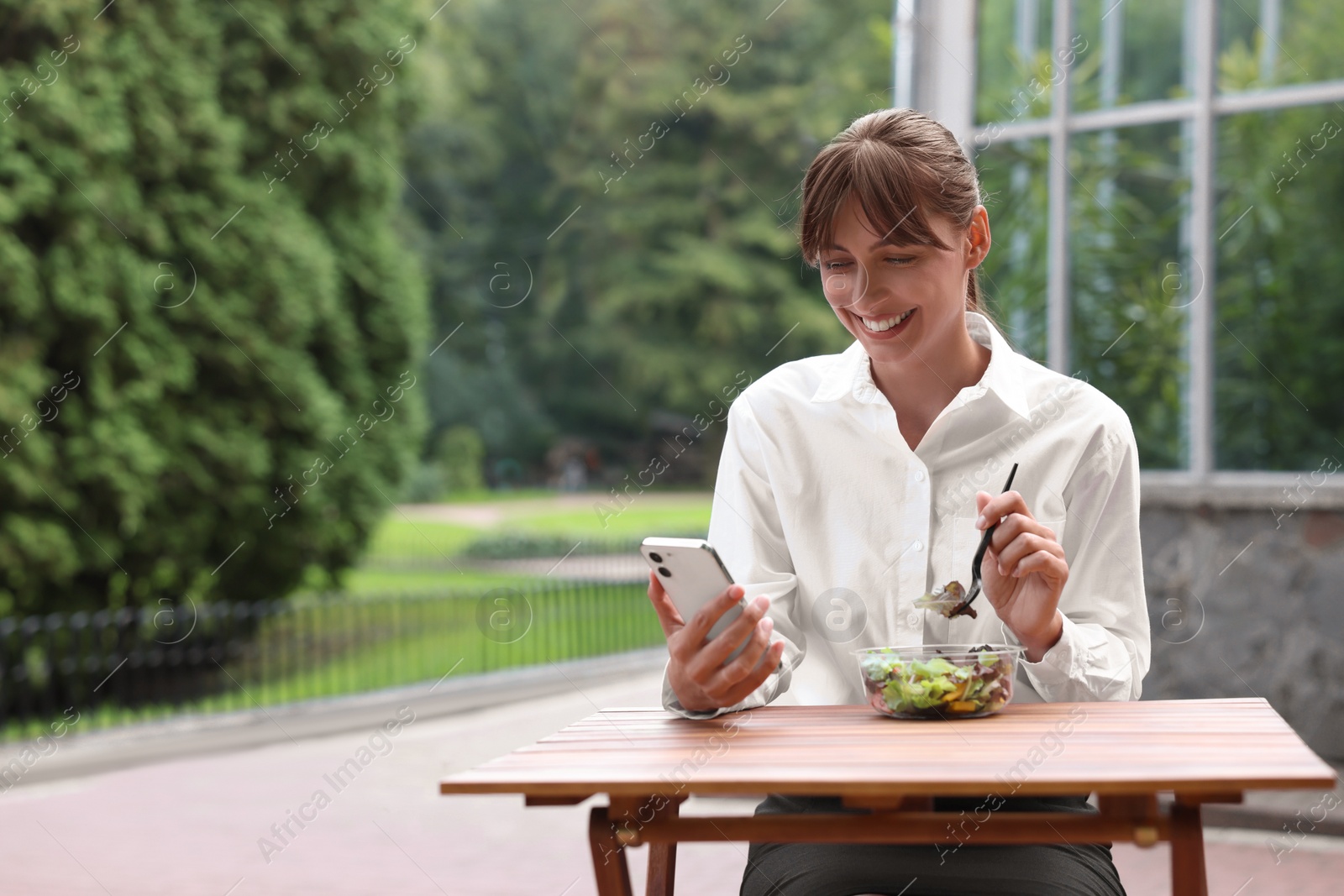Photo of Happy businesswoman using smartphone while having lunch at wooden table outdoors, space for text
