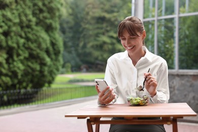 Photo of Happy businesswoman using smartphone while having lunch at wooden table outdoors, space for text