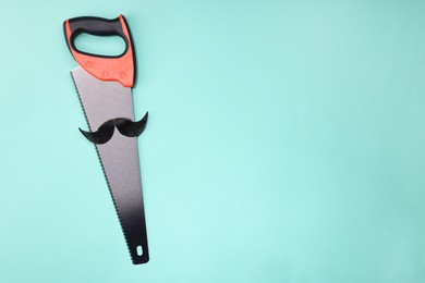 Photo of Man's face made of artificial mustache and hand saw on light blue background, top view. Space for text