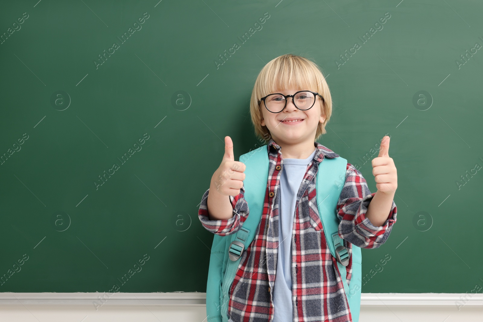 Photo of Happy little school child showing thumbs up near chalkboard. Space for text