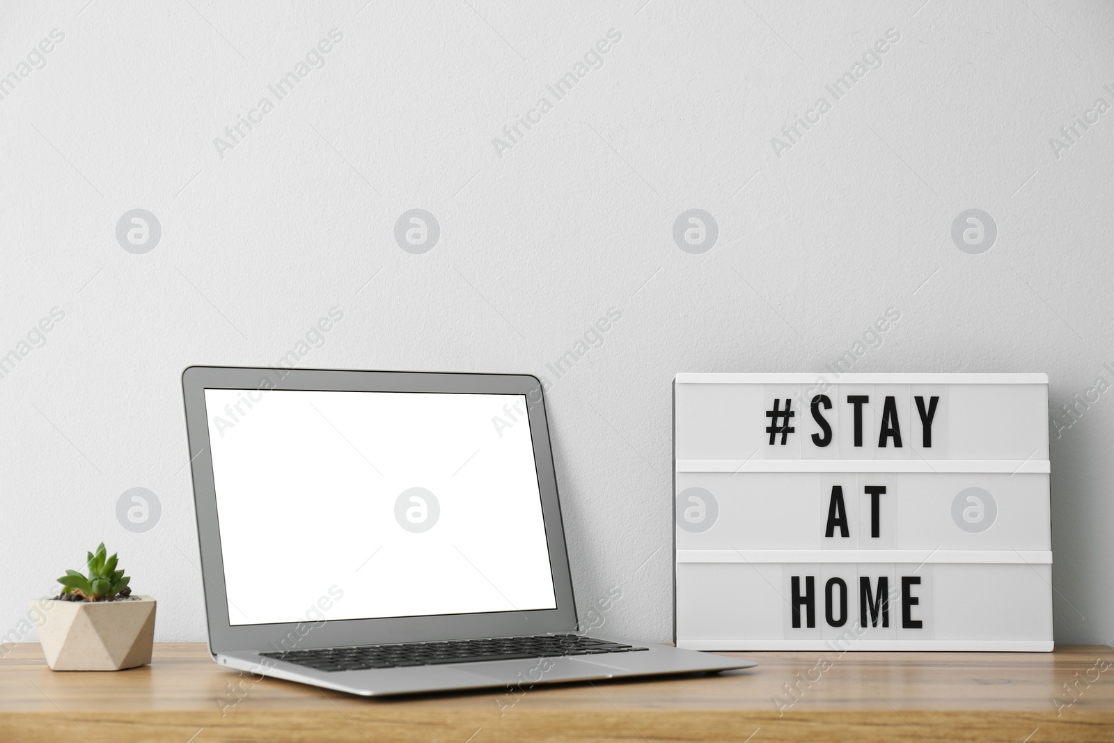 Photo of Laptop and lightbox with hashtag STAY AT HOME on wooden table. Message to promote self-isolation during COVID‑19 pandemic