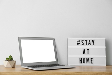 Photo of Laptop and lightbox with hashtag STAY AT HOME on wooden table. Message to promote self-isolation during COVID‑19 pandemic