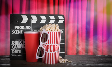 Delicious popcorn, drink, 3D glasses and clapperboard on wooden table against closed red main curtain, space for text