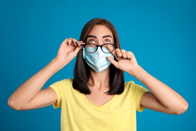 Photo of Young woman wiping foggy glasses caused by wearing disposable mask on blue background. Protective measure during coronavirus pandemic