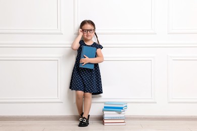 Photo of Cute little girl in glasses with many books near white wall