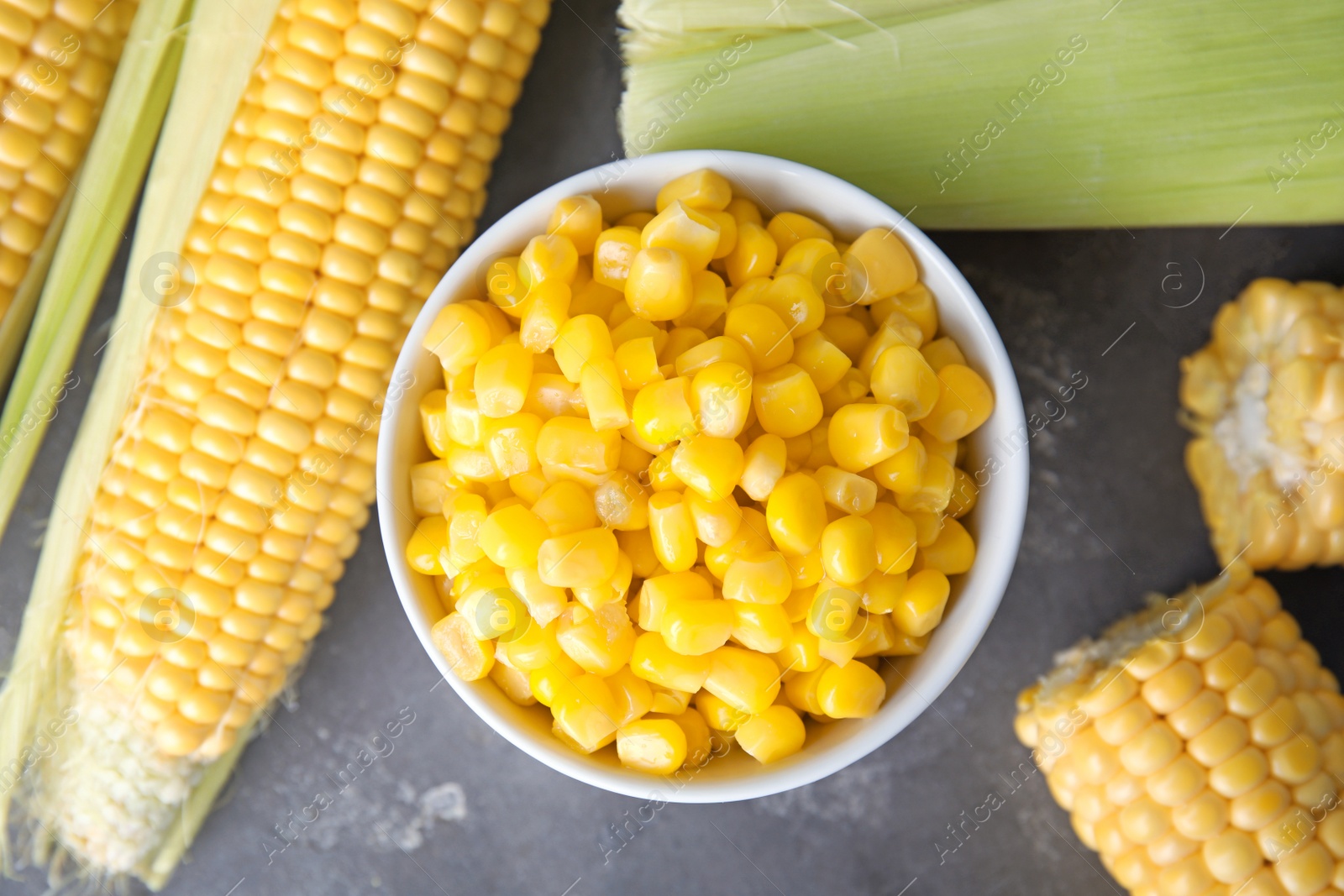 Photo of Bowl with corn kernels and ripe cobs on grey background, top view